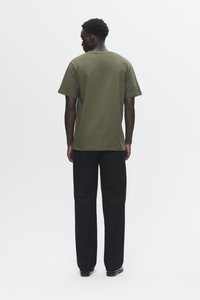 Man standing facing away, wearing an olive green short-sleeve T-shirt, black pants, and black shoes on a white background.