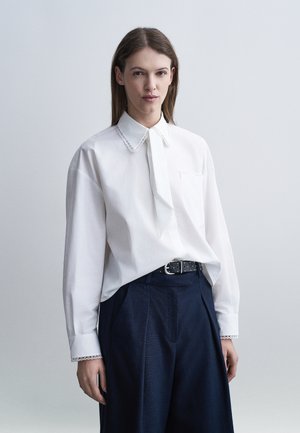 Femme portant un chemisier blanc à manches longues avec bordure en dentelle et ceinture noire, associé à un pantalon taille haute bleu marine, debout devant un fond gris.