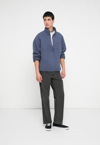 Szary polarowy pulower z półzamkiem, z haftem logo GAP. Para czarno-białych spodni w paski. Czarne trampki. Jednolite tło.