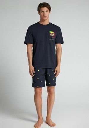 Ung mand iført mørkeblå kortærmet skjorte med citron- og snorkelgrafik og matchende mørkeblå shorts med citronmønster, stående barfodet.