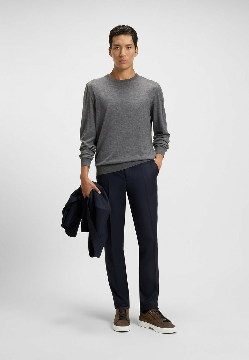 Mann in grauem Pullover, mit marineblauen Hosen und braunen Turnschuhen, hält eine marineblaue Jacke und steht vor einem einfarbigen hellen Hintergrund.