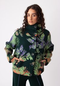 Flisasta jakna v temno zeleni barvi s cvetličnimi vzorci v vijolični, oranžni in svetlo zeleni; ima visok ovratnik, pet gumbov in stranske žepke.