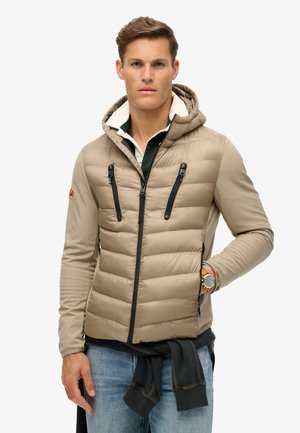 Chaqueta acolchada beige con capucha, con textura acolchada, cremalleras negras y un forro interior en contraste. Combinada con un suéter oscuro y jeans.