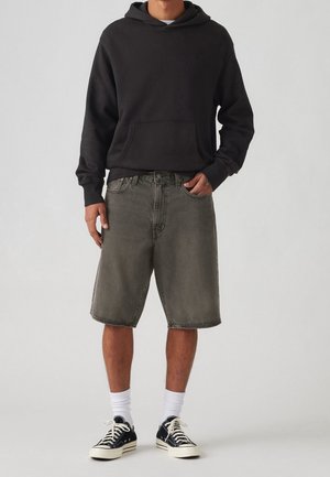 Mand iført sort hættetrøje, grå knælange denimbukser, hvide sokker og sorte sneakers, stående mod en neutral baggrund.