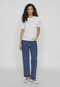 Haut blanc texturé à manches courtes avec des épaules bouffantes, associé à un jean droit en denim bleu et des chaussures plates de couleur claire.