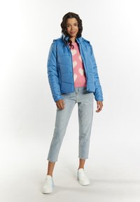 Giacca imbottita blu chiaro con cappuccio, indossata sopra un maglione rosa con motivi a cuore bianchi, abbinata a jeans chiari e scarpe da ginnastica bianche.