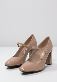 Beige lackläder Mary Jane-klackar, med fyrkantig tå och kraftig klack. Har ett justerbart rem med en guldbuckle.