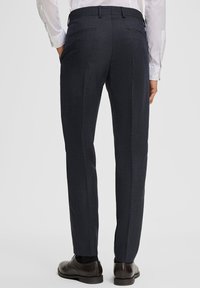 Mann trägt dunkelgraue Anzughose, schwarze Dressschuhe und ein weißes Hemd mit langen Ärmeln, steht mit Händen in den Taschen und hat den Rücken zum Betrachter gewandt.