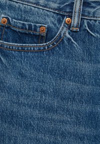 Gros plan sur un jean en denim bleu montrant la poche avant avec rivets en cuivre et coutures détaillées.