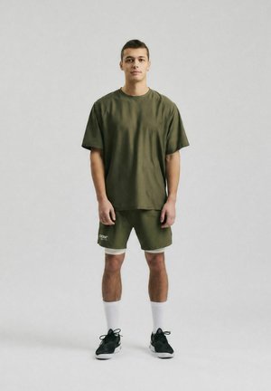 Ung mand står iført olivengrøn løs T-shirt og shorts med hvide sokker og sorte sneakers på en lysegrå baggrund.
