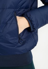 Tmavě modrá prošívaná péřová bunda s kapsou na boku na zip a žebrovanými manžetami pro lepší teplo a přizpůsobení.