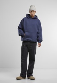 Marineblå oversized hettegenser med lommer foran, kombinert med mørke bukser og beige slip-on sko. Modellen har på seg en lys grå strikket lue.