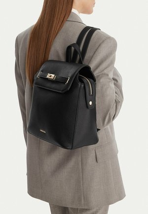 Frau in einem grauen Anzug trägt einen schwarzen Lederrucksack von Lasocki mit goldenen Beschlägen auf dem Rücken und ist von der Kamera weggewandt.