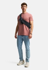 Camiseta de manga corta rosa para hombre, jeans de mezclilla azul, suéter gris colgado sobre el hombro, zapatillas beige, de pie contra un fondo neutro.