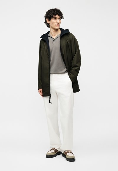 Jeune homme portant une veste à capuche vert foncé, un polo gris, un pantalon blanc et des mocassins beiges à semelles foncées, debout avec une main dans la poche.