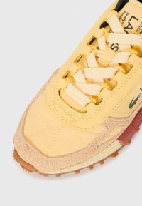 Yellow and beige sneaker with textured suede toe cap, thick laces, and printed text on the tongue and side.