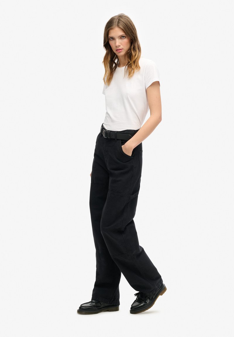 Schwarze weit geschnittene Hose mit Seitentaschen, kombiniert mit einem enganliegenden weißen T-Shirt. Die Schuhe sind schwarz mit glänzender Oberfläche. Einfaches, lässiges Design.