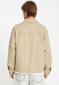 Chaqueta grande beige con cuello de muesca, puños abotonados y una textura suave. Lleva puesta sobre una camisa blanca y combinada con pantalones gris claro.