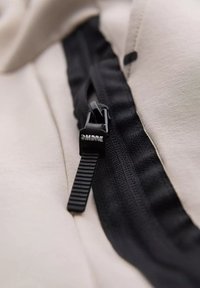 Světlý béžový materiál s černým zipem. Zip má texturovanou táhlo, na kterém je stříbrným písmem napsáno "OMBRE". Detailní pohled.