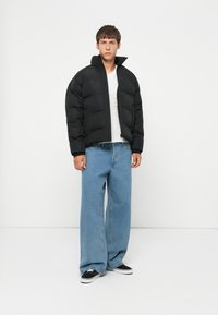 Ung mann iført en svart boblejakke, hvit skjorte, vide blå jeans og svarte joggesko som står mot en hvit bakgrunn.