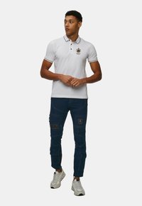 Polo bianco con colletto e logo neri a contrasto, abbinato a pantaloni cargo blu con tasche a toppa e tessuto texture, indossato con sneakers bianche.