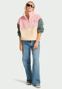 Sudadera de forro polar con media cremallera en rosa, crema y verde; presenta un cuello, textura suave y un logo; combinada con jeans acampanados azules.