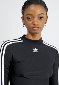 Blusa preta de mangas longas com gola alta, logótipo branco da Adidas no peito e riscas brancas a correr pelas mangas. Tecido elástico, design ajustado.