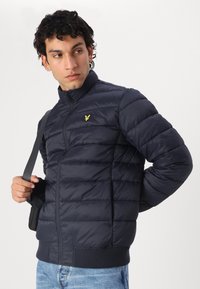 Marineblå puffjakke med vertikal quilt, høy krage og elastiske ermer. Har en liten gul logo på brystet og glidelås-lukking.