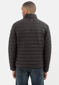 Sort vinterjakke i pufferstil med lodrette syninger, høj krave og et lille mærke på bagsiden. Bæres med blå jeans, der viser et glat, let materiale.