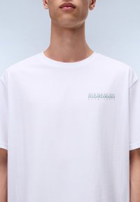Vit bomulls-T-shirt med rund halsringning, med ett subtilt ljusgrönt tryck av "NAPAPIJRI geographic" på vänster bröstrygg.