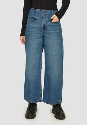 Jean large en denim bleu moyen, taille haute, avec deux poches avant, fermeture à bouton et ourlet effiloché, associé à des bottes noires à lacets.