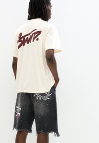 T-shirt in cotone beige con logo bordeaux sulla schiena; abbinato a pantaloni corti in denim neri con accenti grafici bianchi e orli sfrangiati.