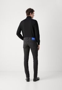 Black long-sleeve shirt, slim-fit black jeans with blue 'HUGO' patch on waistband, worn with black shoes, viewed from the back.