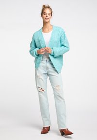 Hellblauer, grob gestrickter Cardigan mit V-Ausschnitt und oversized Ärmeln, kombiniert mit ausgebleichten, lässigen Jeans und braunen, gehäkelten Mules.