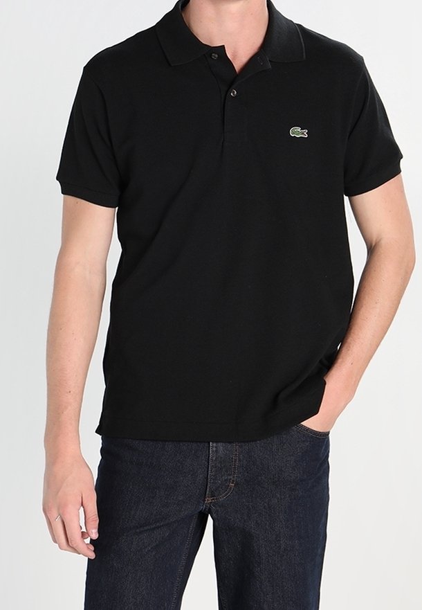 Homme portant un polo noir à manches courtes avec un logo crocodile vert et un jean bleu foncé, une main dans la poche.