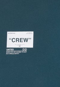 Camiseta de algodón color teal con cuello redondo que presenta una etiqueta de tela blanca con el texto "CREW" y "UNITED" impreso en negro, centrado en el pecho.