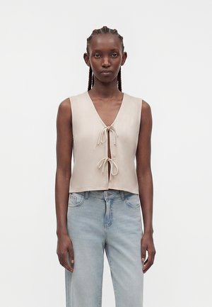 Γυναίκα με πλεξούδες, που φοράει μια μπεζ αμάνικη μπλούζα δεμένη με δύο φιόγκους εμπρός και ανοιχτό μπλε ψηλόμεσα τζιν, στέκεται μπροστά σε λευκό φόντο.