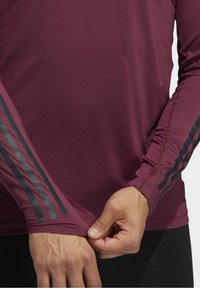 Długi rękaw koszulka sportowa w głębokim burgundzie, z czarnymi paskami na rękawach, gładką fakturą materiału i dopasowanymi mankietami.