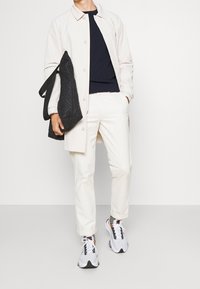 Trench-coat beige clair, pull en maille bleu marine, pantalon blanc fuselé et chaussures de sport grises. Sac matelassé noir tenu dans la main gauche.