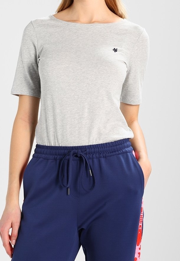 Femme portant un haut gris clair à manches courtes avec un petit logo noir et un pantalon bleu marine à cordon avec des bandes rouges sur les côtés.