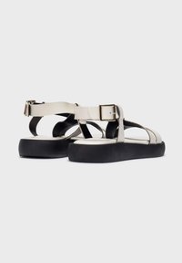 Sandalias beige claras con una suela negra texturizada. Presenta correas cruzadas y un acento de hebilla dorada. Diseño plano adecuado para el uso casual.