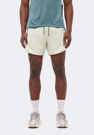 Modèle masculin portant un short de sport beige clair avec cordon, des chaussettes blanches et des chaussures de course grises, debout devant un fond uni.