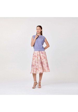 Femme portant un haut en maille lavande sans manches, une jupe midi florale rose et des sandales blanches à talons hauts, debout devant un fond uni.