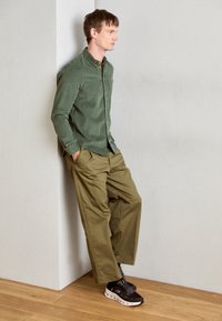 Camicia a bottoni verde in corduroy, pantaloni larghi color oliva e scarpe da ginnastica nere con suole bianche. Uomo appoggiato a un muro.
