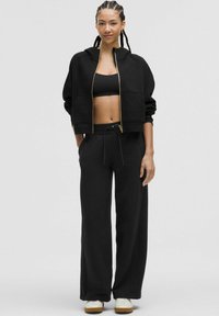 Sudadera corta negra con cremallera dorada, combinada con pantalones negros holgados. Tela suave y lisa, con un corte relajado y cintura con cordón.