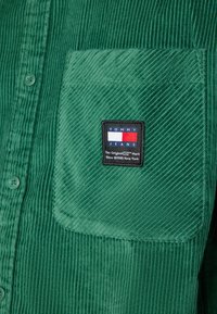 Zelená manšestrová košile s knoflíkovým zapínáním, vybavená levým kapsičkou na prsou. Obsahuje obdélníkovou nášivku s logem Tommy Jeans. Pruhovaná textura.