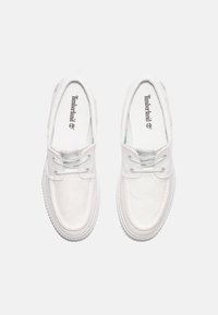 White canvas slip-on shoes with silver eyelets and laces. Textured sole, Timberland branding on the insole, and a minimalist design.