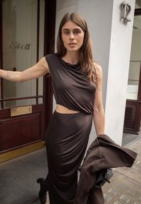 Brahno oblečeno krilo z asymetričnim izrezom, teksturirana tkanina in brezrokavni dizajn. Model drži rjav plašč in stoji ob vhodu v trgovino.