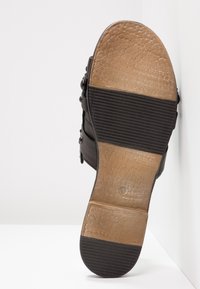 Semelle de chaussure en cuir avec des grips en caoutchouc texturés au niveau des orteils, du talon et du milieu du pied, placée contre une surface blanche et un angle de mur.