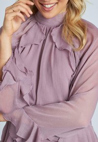 Blusa lilac morbida con colletto alto, dettagli a rouches sovrapposti e trama trasparente. Maniche lunghe con polsini fru-fru, accenti di cuciture delicate.